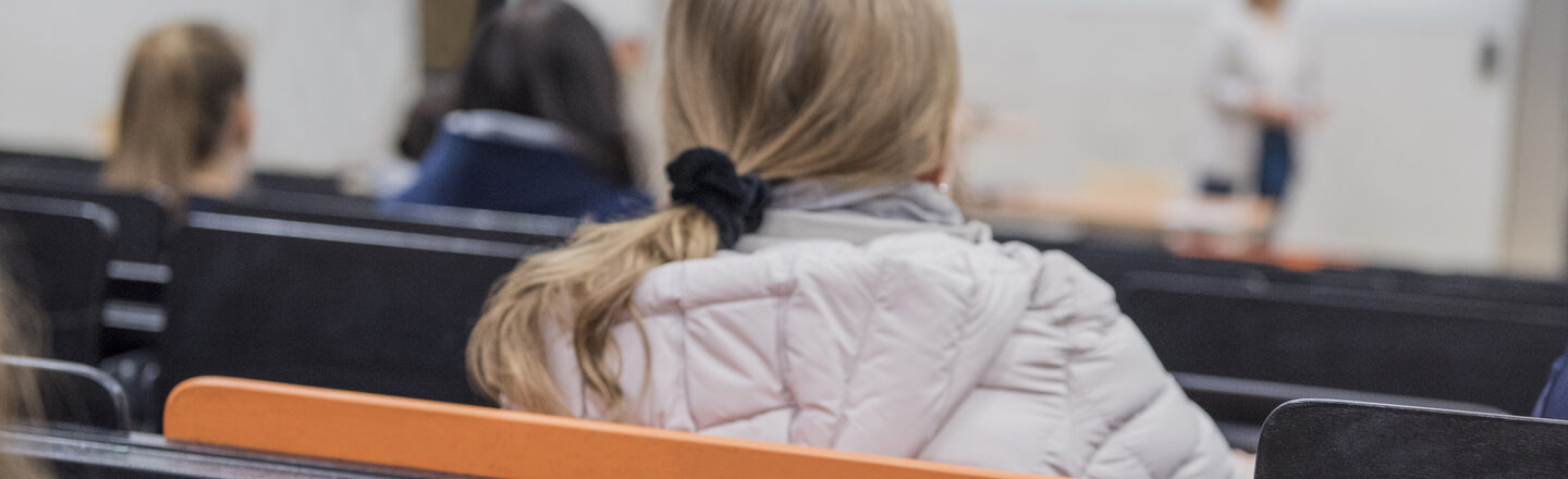 Photo of a young woman in the back of a lecture hall row, looking towards the lecturer. In the row behind her, the table has been opened and printed materials are lying on it. __ <br>Young woman in a row of lecture halls, looks forward to the speaker. In the row behind it, the table has been opened, and there are print materials on it.