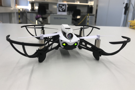 Eine Parrot Mambo Drohne auf einem Tisch im Labor stehend mit Blick zur Kamera. Zwei LED-Lämpchen vorne, die aussehen wie die Augen der Drohne. Oben auf der Drohne ist ein Kamaramodul montiert.