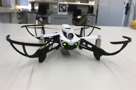 A Parrot Mambo drone standing on a table in the laboratory facing the camera. Two LED lights at the front that look like the eyes of the drone. A camera module is mounted on top of the drone.