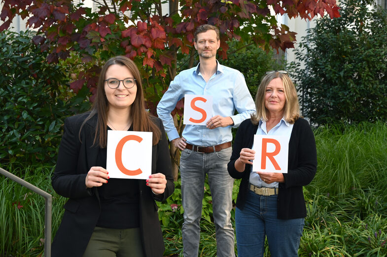 Drei Personen halten jeweils ein Blatt Papier mit je einem orangefarbenen Buchstaben hoch. Von links nach rechts liest man so CSR.