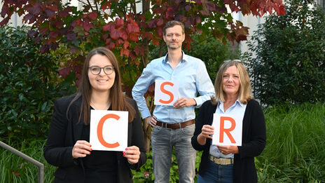 Gruppenbild CSR-Office-Teams: Drei Personen halten jeweils ein Blatt Papier mit je einem orangefarbenen Buchstaben hoch. Von links nach rechts liest man so CSR.