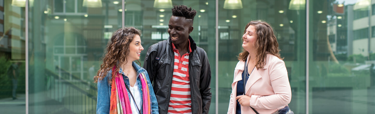 Photo of two female and one male students standing side by side in front of the canteen building, they laugh at each other. They laugh at each other.__Two female and one male students stand side by side in front of the canteen building, they laugh at each other.