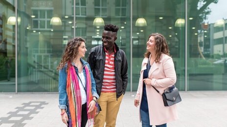 Foto von zwei Studentinnen und einem Studenten, die nebeneinander vor dem Mensagebäude stehen. Sie lachen einander an.__Two female and one male students stand side by side in front of the canteen building, they laugh at each other.