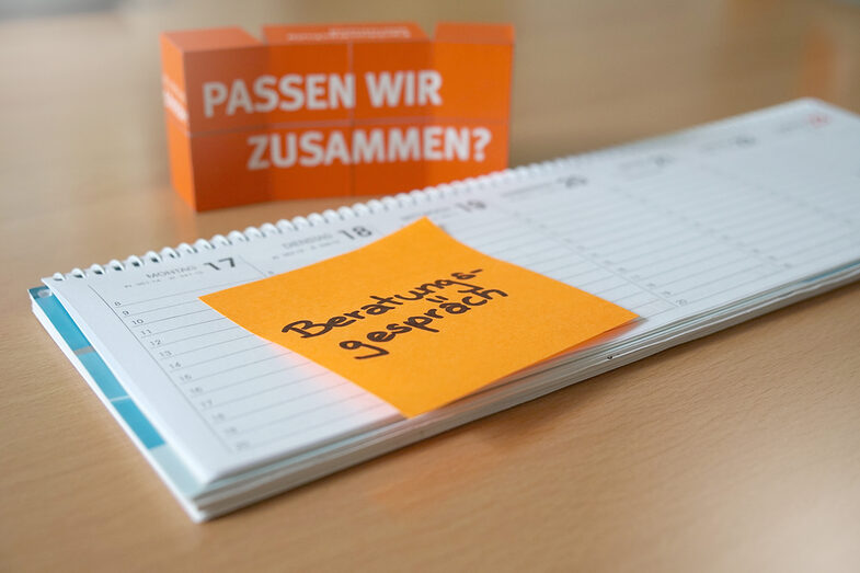 Foto eines Tischkalenders, auf dem ein orangefarbenes Post-It mit der Notiz „Beratungsgespräch“ klebt. Im Hintergrund stehen gestapelte Würfel, auf denen „Passen wir zusammen?“ steht.