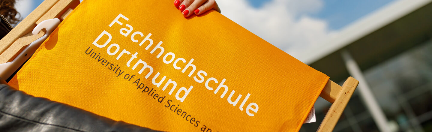 Photo of a female student - strongly cropped. She wears an orange folding chair with the FH logo. __ Female student - strong in the cut - wears an orange folding chair with the FH logo.