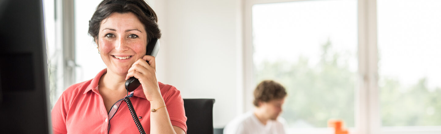 Photo of an employee sitting at her desk talking on the phone and smiling at the camera. In the background, a man is working on his laptop.