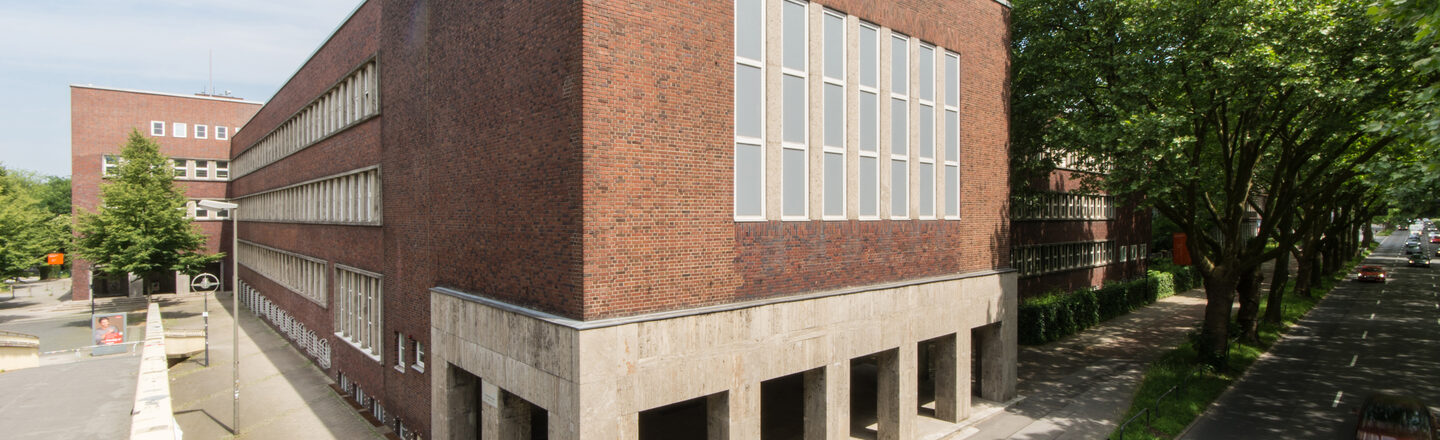 Photo with a view of parts of a building of the Fachhochschule Dortmund at Max-Ophüls-Platz with forecourt and sidewalk as well as street with cars next to it.