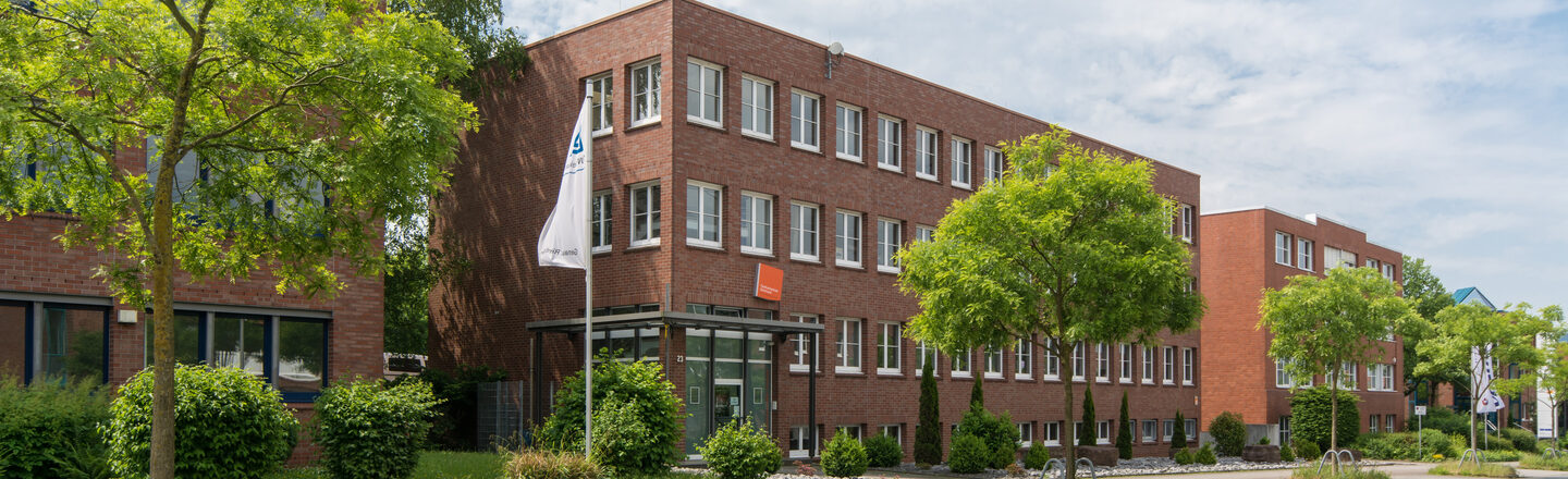 Photo of the Fachhochschule Dortmund site in Otto-Hahn-Straße from the outside. It is a red brick building. In the foreground are a road and some trees.