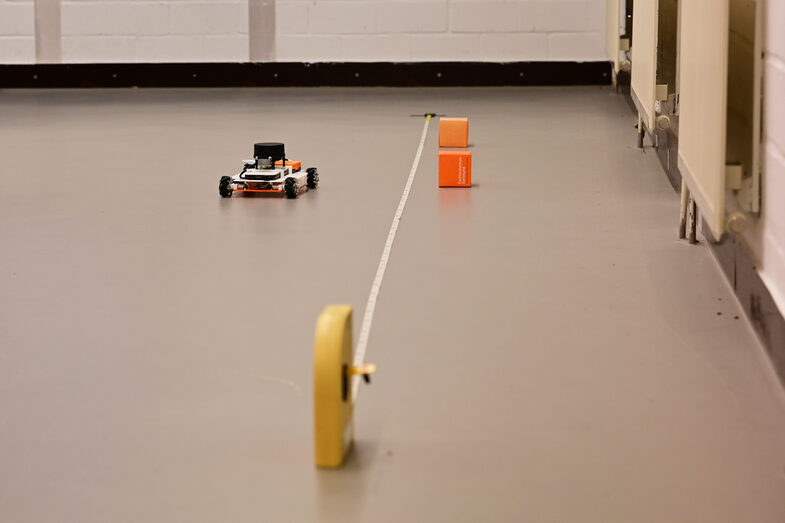 EduRob stands on the floor of the laboratory to the left of a measuring tape. Small FH cubes are placed at marker distance points to mark the distance.