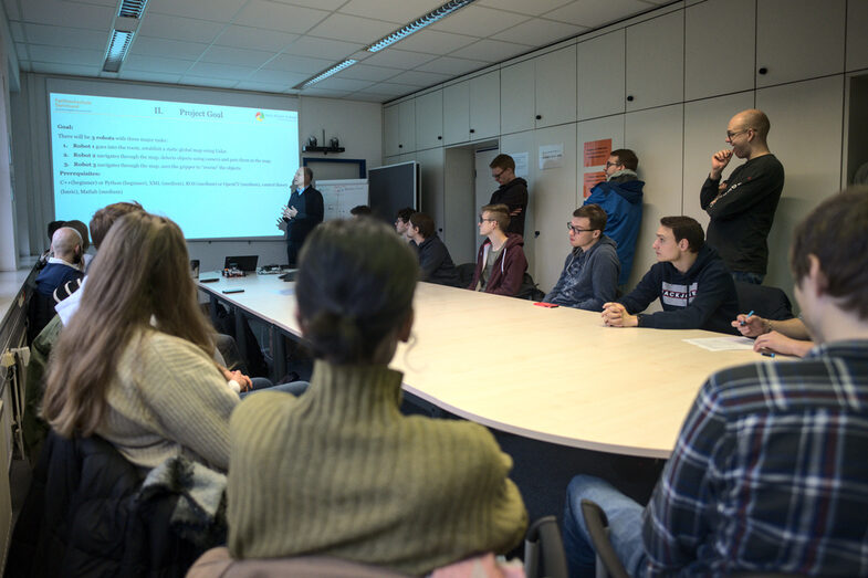 Students at a large table in the IDiAL meeting room. Prof. Dr. Christof Röhrig stands at the screen and explains the objectives of the block week using a projected slide.