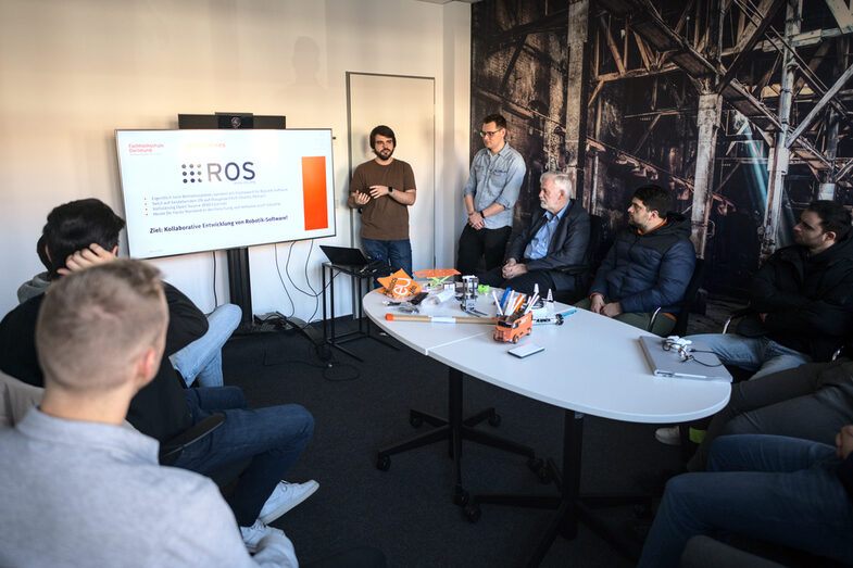 Students sit around a table in the meeting room. On the table are various grippers for robots. At the end of the table, Alexander Miller is standing in front of a TV screen showing a slide about ROS and giving a lecture.