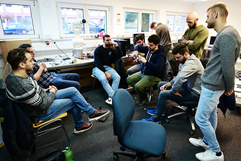 Die Studierenden des Teams Greifer haben sich in einem Stuhlkreis im Automatisierungstechnik-Labor des IDiALs zusammengefunden. In der Mitte setzt ein Student den Greifer zusammen. Alle Teilnehmer beobachten dies interessiert.