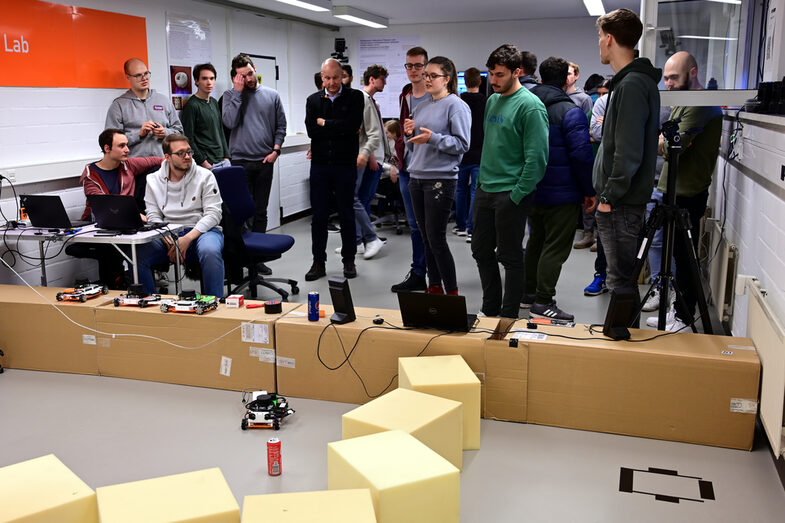 The participants of the block week are gathered in the robot lab in the background behind the scenario that has been set up. A student explains the implemented vision system. In the foreground you can see the set-up scenario, consisting of foam cubes and cardboard boxes. An EduRob with a stereo camera and a drinks can can be seen in the middle of the scenario.
