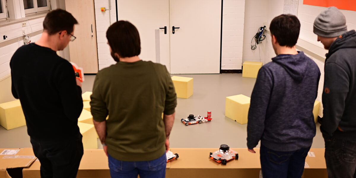Vier Personen (von hinten zu sehen) stehen vor dem aufgebauten Szenario im Roboterlabor. Ein EduRob greift eine Getränkedose mittels des während der Blockwoche entwickelten Greifers. Zwei Personen diskutieren über einen EduRob, den die linke Person in den Händen hält.