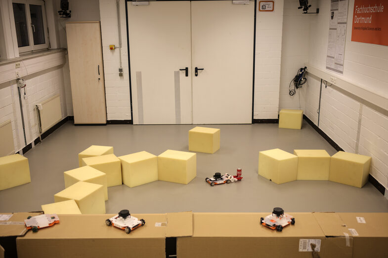 Aufnahme der Deckenkamera des Robotik-Labors des IDiALs. Zu sehen sind Kartons und Schaumstoffwürfel, die einen Parcours für die Roboter der studentischen Teilnehmer der Robotik-Blockwoche formen. In mitten des Szenarios findet sich ein EduRob (kleiner mobile Roboter) und eine Getränkedose.