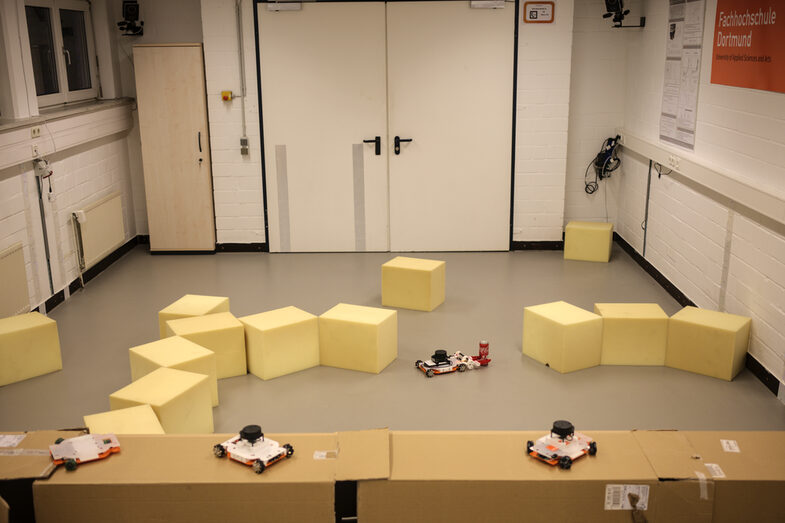 Image from the ceiling camera in the IDiAL robotics lab. You can see cardboard boxes and foam cubes that form a course for the robots of the student participants of the robotics block week. In the middle of the scenario is an EduRob (small mobile robot) and a drinks can.