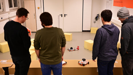 Vier Personen (von hinten zu sehen) stehen vor dem aufgebauten Szenario im Roboterlabor. Ein EduRob greift eine Getränkedose mittels des während der Blockwoche entwickelten Greifers. Zwei Personen diskutieren über einen EduRob, den die linke Person in den Händen hält.
