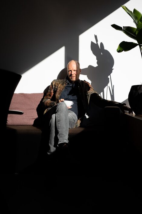 A person with a cup in his hand sits on a couch. From the right, bright daylight cuts into the shadowy room and casts the hard shadow of a plant on the back wall.