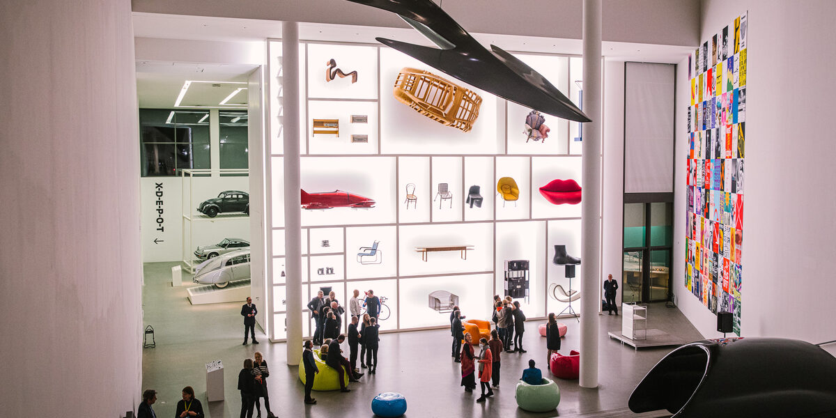 A high room in a museum with around two dozen people standing in it. Works of art hang on the walls and from the ceiling.