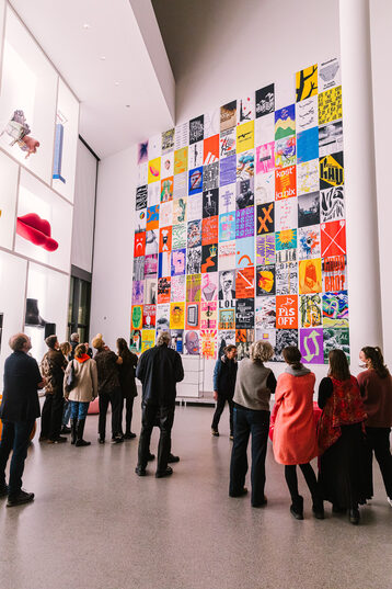 Several people stand in a museum room with their backs to the camera, looking at a high wall covered with posters.
