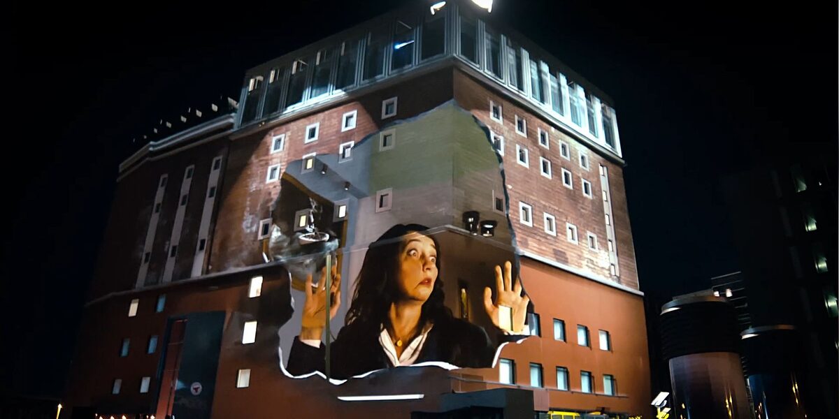 Projection onto the façade of the Dortmunder U: A woman presses her face against an invisible pane of glass in surprise.