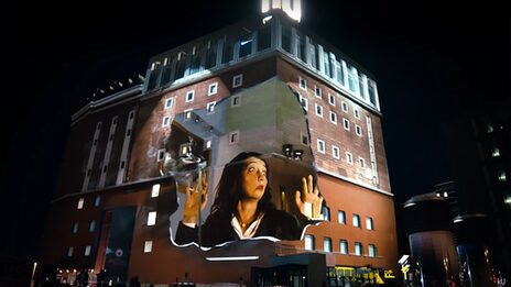 Projektion auf die Fassade des Dortmunder U: Eine Frau drückt ihr Gesicht überrascht gegen eine nicht sichtbare Glasscheibe.