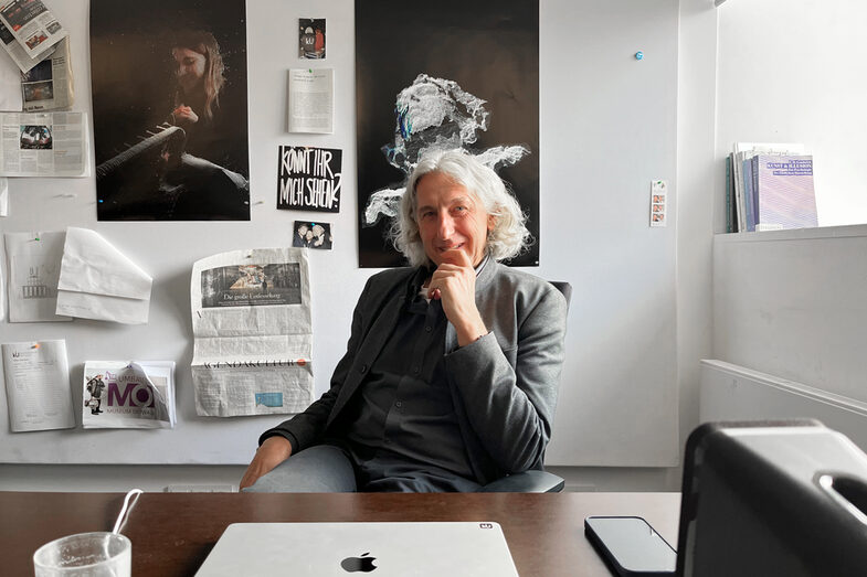 A man sits at a desk, posters and newspaper clippings hang on the wall behind him, daylight falls in from the right.