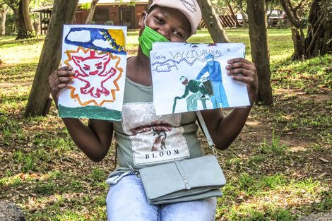 A girl holds up two pictures painted by children. The pictures focus on the coronavirus pandemic and the topic of vaccination.