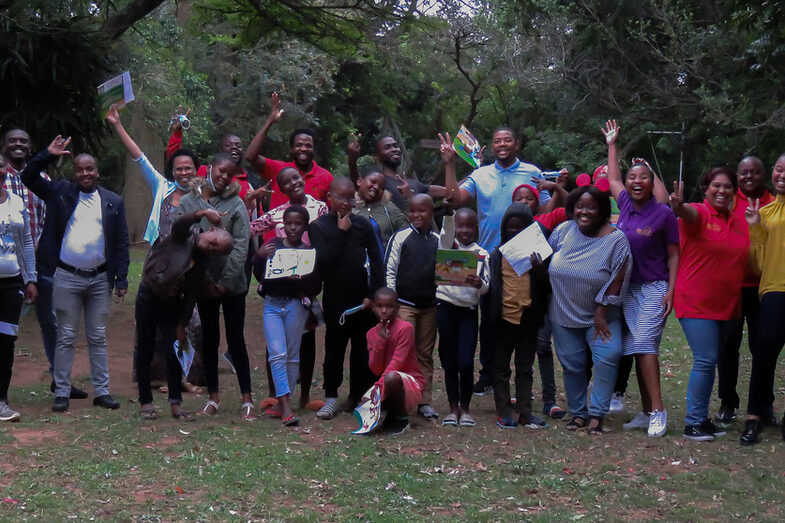 A large group of people stand outside under the trees for a group photo. They are adults and children, cheering.