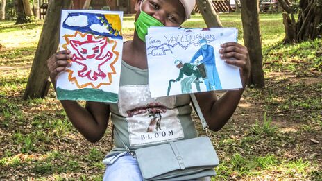 Ein Mädchen hält zwei von Kindern gemalte Bilder hoch. Die Bilder thematisieren die Corona-Pandemie und auch das Thema Impfung.