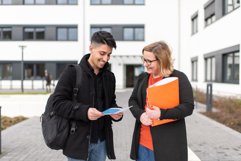 Photo of an employee and a student standing in front of a campus building. She is holding an orange folder under her arm, he has a flyer in his hand. They both look at the flyer and laugh.