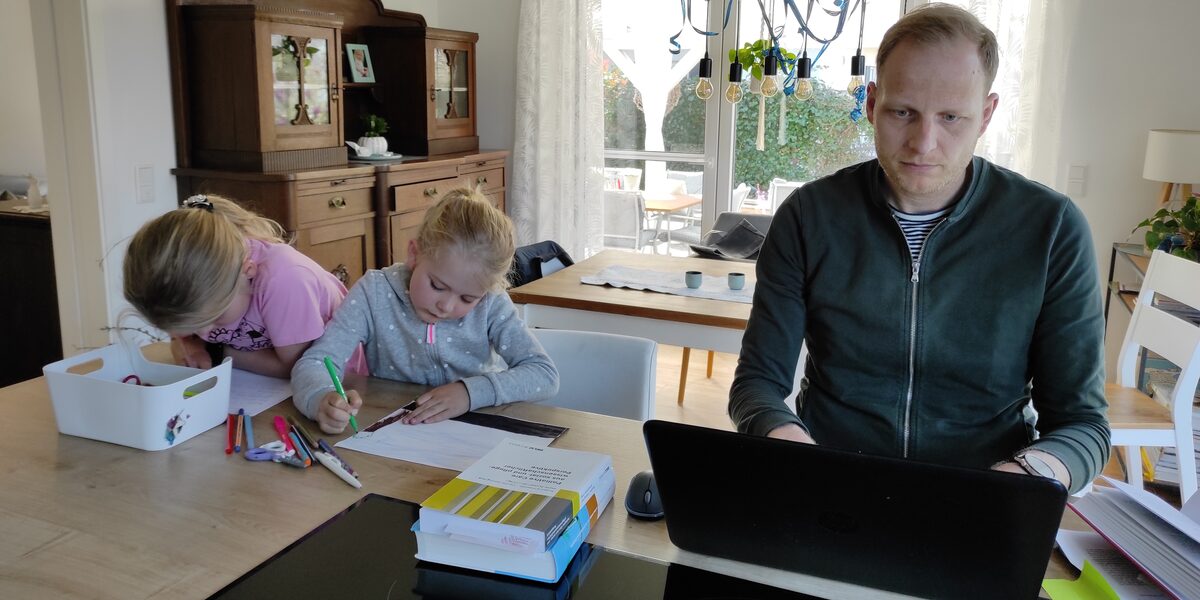 Der Stipendiat Jens Terbrack sitzt am Tisch und arbeitet an seinem Laptop. Daneben malen seine beiden Töchter Bilder.
