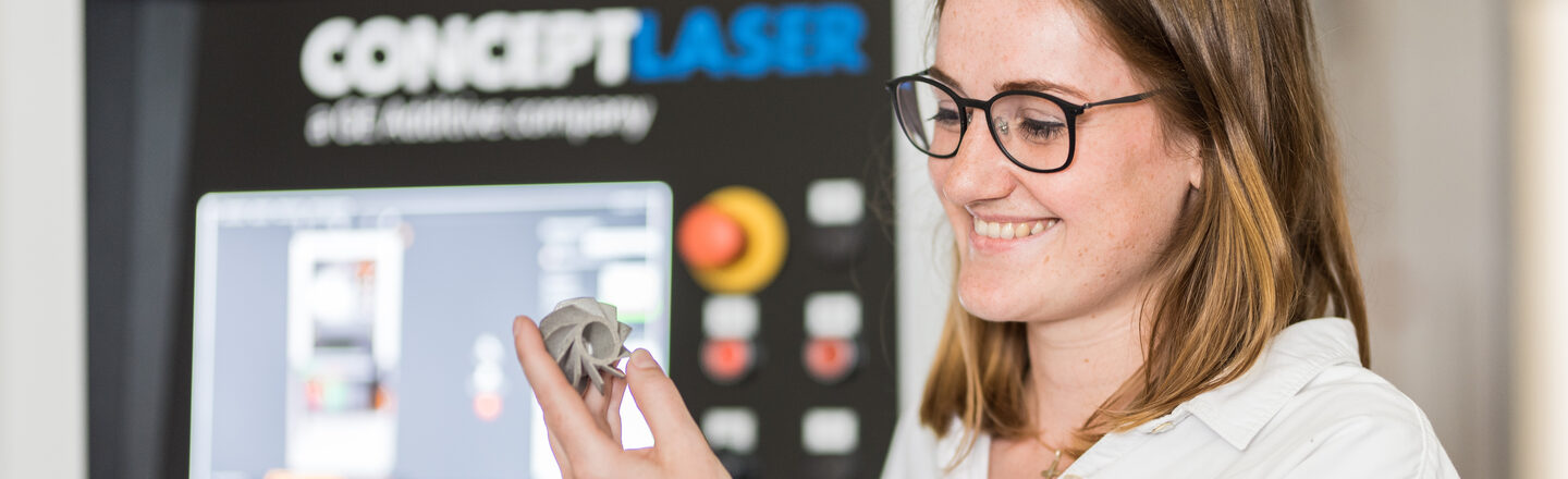 Foto von einer Frau, die sich ein 3D gedrucktes Metallmodell in ihrer Hand anguckt, im Hintergrund unscharf ist der Drucker erkennbar.