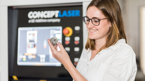 Foto von einer Frau, die sich ein 3D gedrucktes Metallmodell in ihrer Hand anguckt, im Hintergrund unscharf ist der Drucker erkennbar.