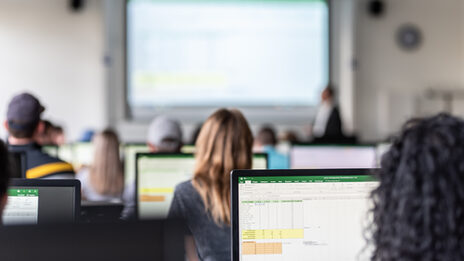 View from behind into the computer room with students, some Excel spreadsheets are visible on the monitors.