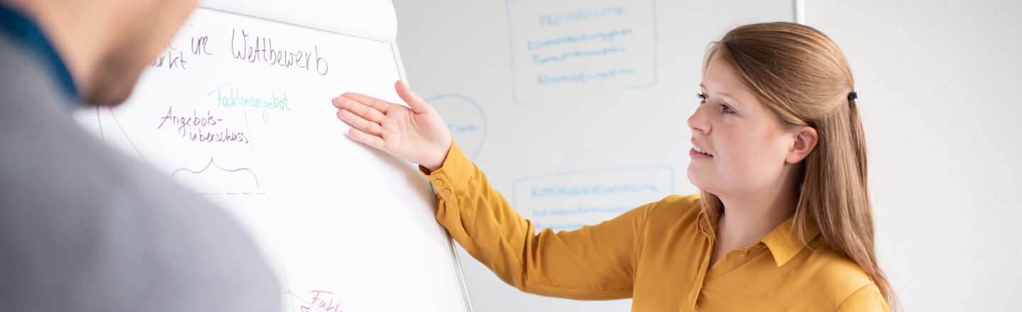 Studentin zeigt auf das Wort "Wettbewerb" auf einem Flipchart und erläutert ihrem Kommilitonen gegenüber etwas.