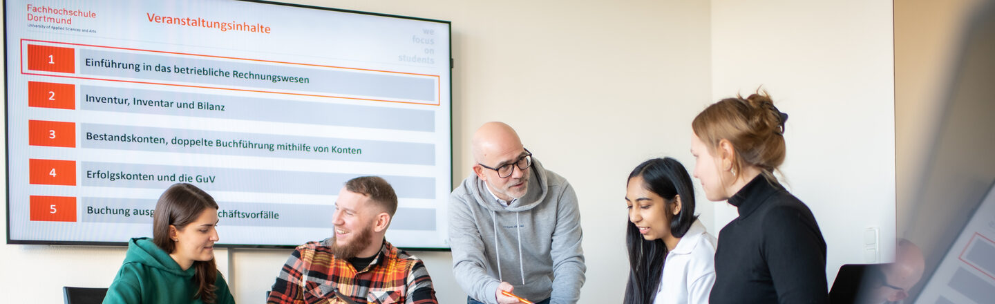 Photo of several students sitting at a table. A lecturer is standing in the middle of them, leaning down and explaining something.