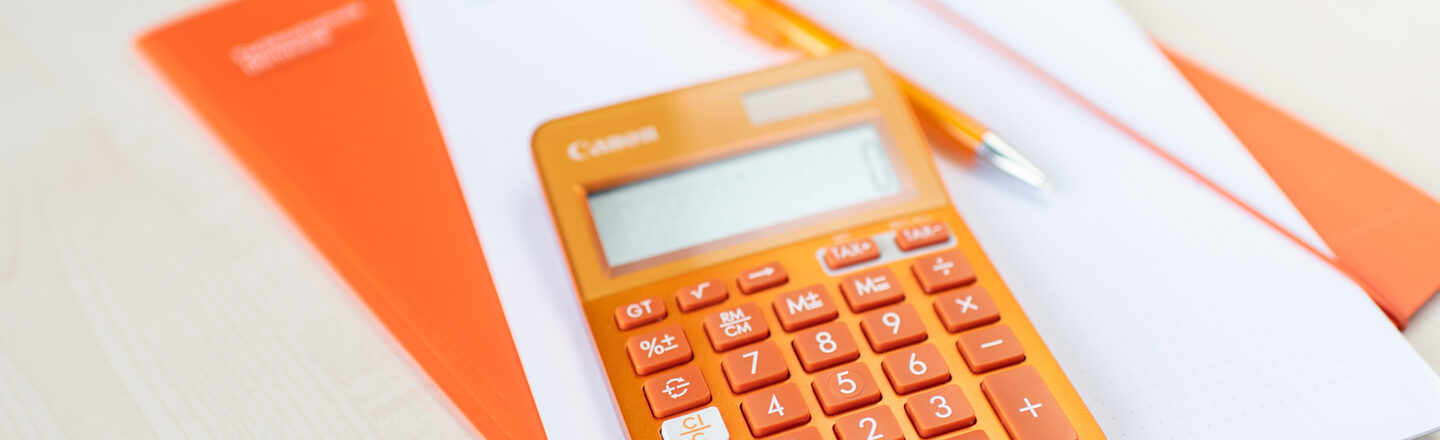Ein orangener Taschenrechner liegt auf einer orangenen Mappe und einem Stapel Zettel.