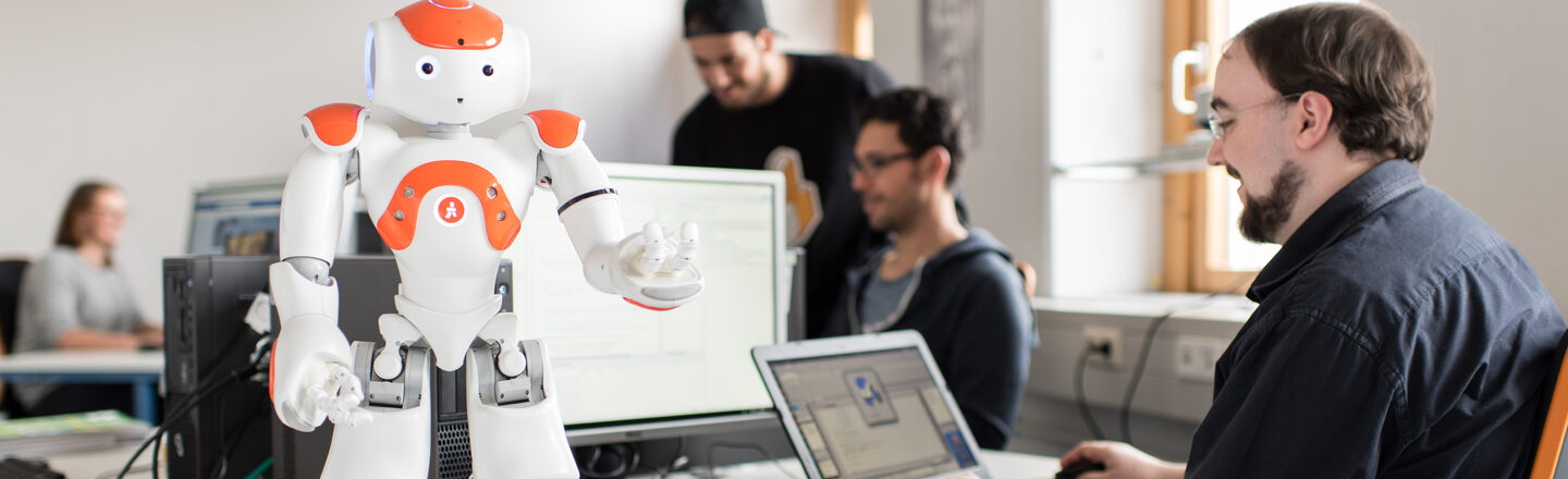 Photo of the NAO robot Eva standing on the table in front, arms raised. Next to it, a student sits at the laptop to give Eva commands.