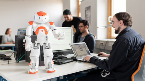 Foto vom NAO-Roboter Eva, der vorne auf dem Tisch steht, Arme gehoben. Daneben sitzt ein Student am Laptop, um Eva Befehle zu geben.