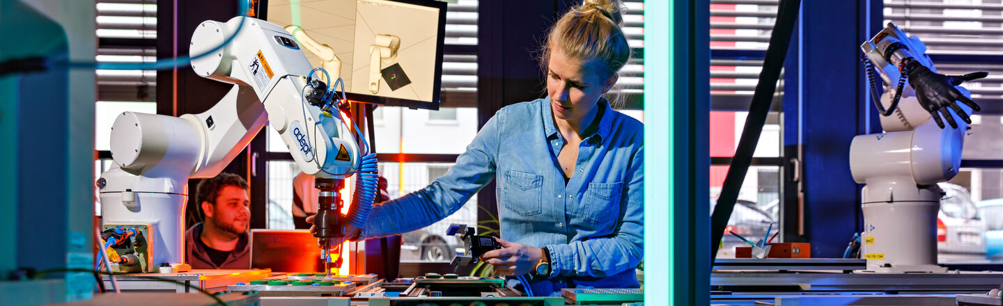 Foto von einem Förderband mit Roboterarmen, hinter dem eine Mitarbeiterin steht, die mit der rechten Hand zu einem Roboterarm greift. __ Behind a conveyor belt with robot arms stands an employee who reaches for a robot arm with her right hand.