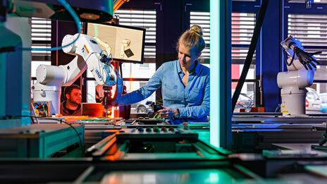 Foto von einem Förderband mit Roboterarmen, hinter dem eine Mitarbeiterin steht, die mit der rechten Hand zu einem Roboterarm greift. __ Behind a conveyor belt with robot arms stands an employee who reaches for a robot arm with her right hand.