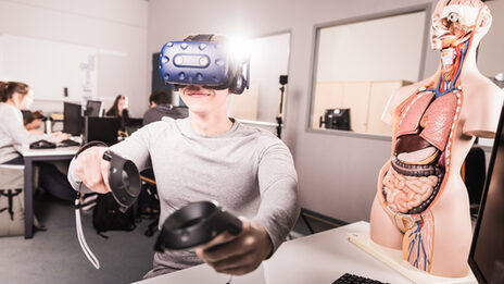 Foto von einem Studenten, der eine VR-Brille auf hat und Controller in der Hand hält, er sitzt an einem Rechnerarbeitsplatz.__A student has VR glasses on and controller in his hands,  is sitting at a computer workstation.