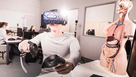 Photo of a student who has VR glasses on and controller in his hands, is sitting at a computer workstation __A student has VR glasses on and controller in his hands, is sitting at a computer workstation.