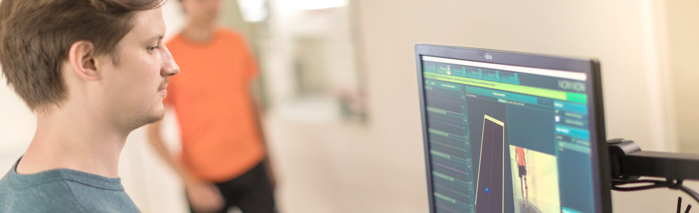 Photo of a young man standing at a PC and carrying out a motion analysis for another man in the background who is walking on a pressure measuring plate in sportswear.