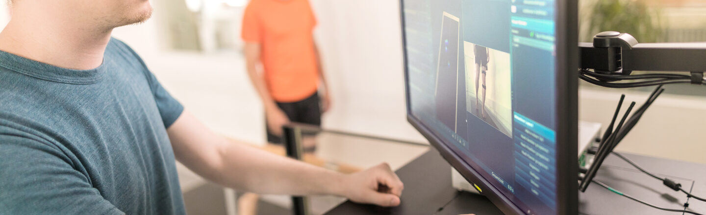 Photo of a young man standing at a PC and carrying out a motion analysis for another man in the background who is walking on a pressure measuring plate in sportswear.