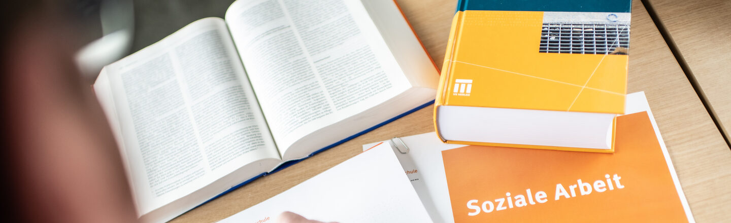 Photo of someone writing something on a pad with an orange pen. Next to it are books and a notebook labeled "social work".