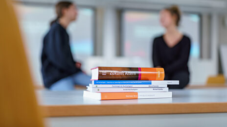 Foto eines Bücherstapels auf dem Tisch. Im Hintergrund unscharf zu erkennen zwei Personen, die sich unterhalten.