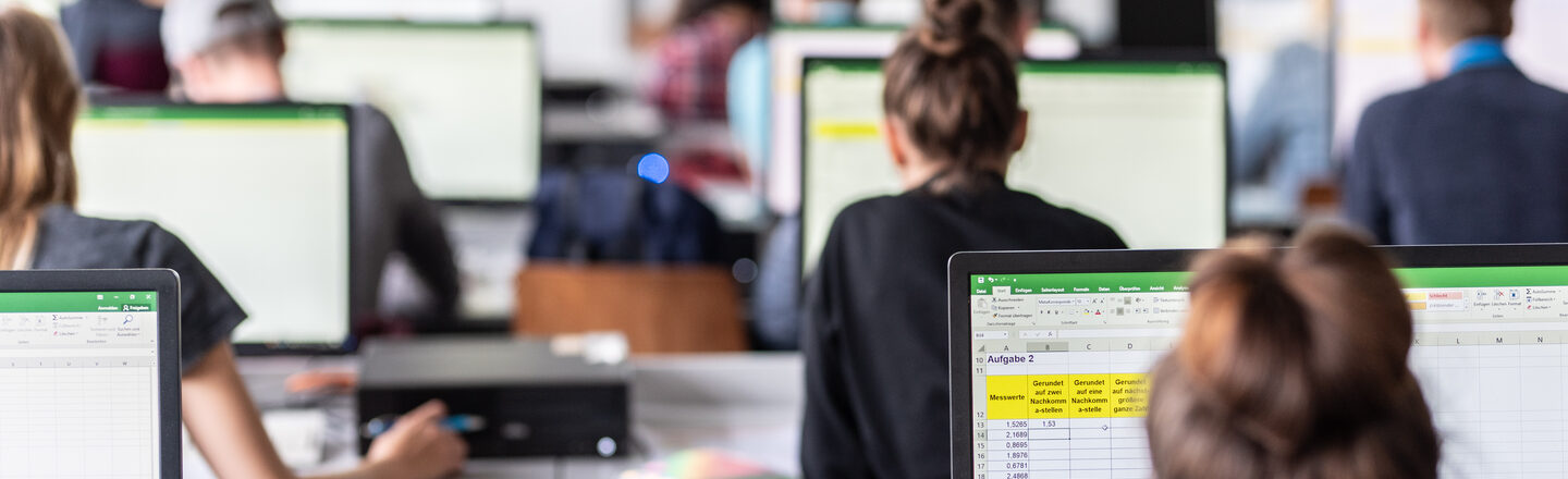 View from behind into the computer room with students, some Excel spreadsheets are visible on the monitors.