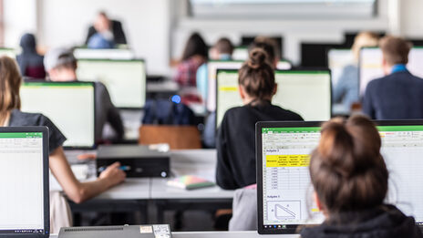 View from behind into the computer room with students, some Excel spreadsheets are visible on the monitors.